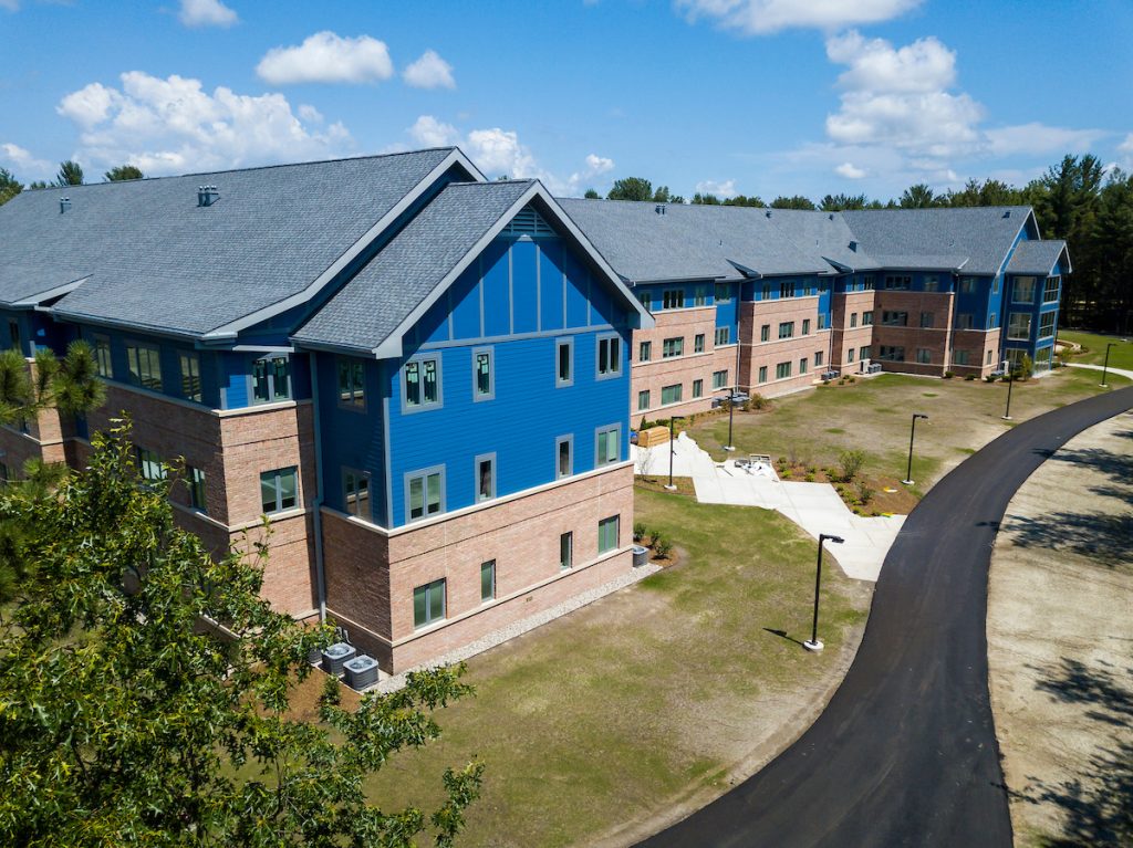 Aerial view of the North Village Apartments, a modern, multi-story blue and brick building with a grey roof, surrounded by a lightly landscaped area and a paved driveway on a sunny day with scattered clouds.