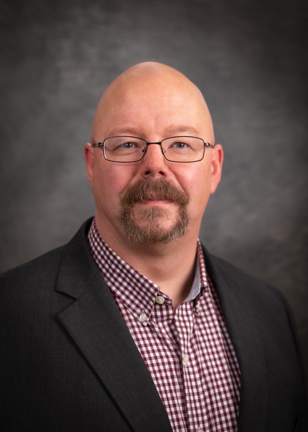 A man with glasses, a bald head, and a goatee, wearing a checkered shirt and a dark blazer, standing against a gray background.