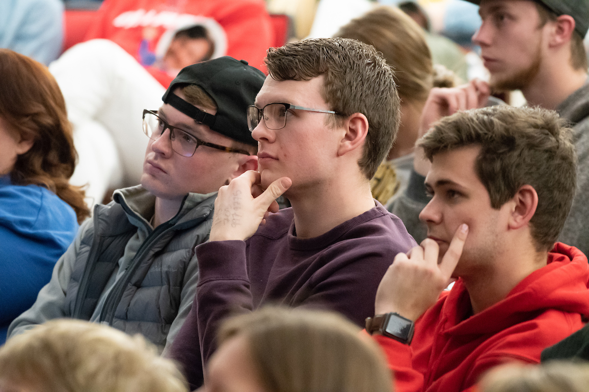 Three young men seated among a group of people, attentively listening during their visit to campus, with focused expressions. One wears a black cap and glasses, the second wears glasses and a maroon sweater, and the third sports a red hoodie.