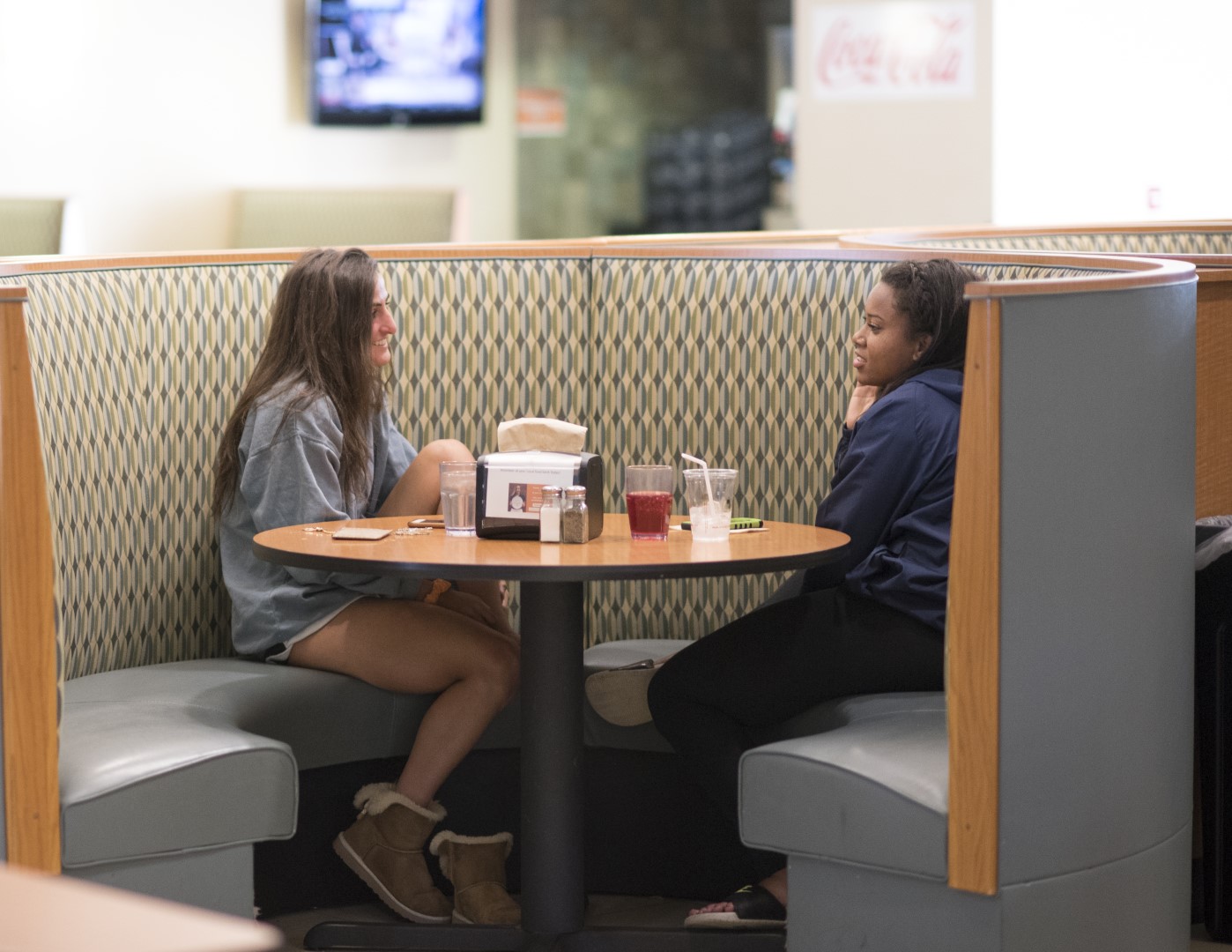 Two people sit in a restaurant booth, engaged in conversation about their recent visit to campus. They are facing each other across a round table with drinks, napkins, and condiments on it. A TV is visible in the background.