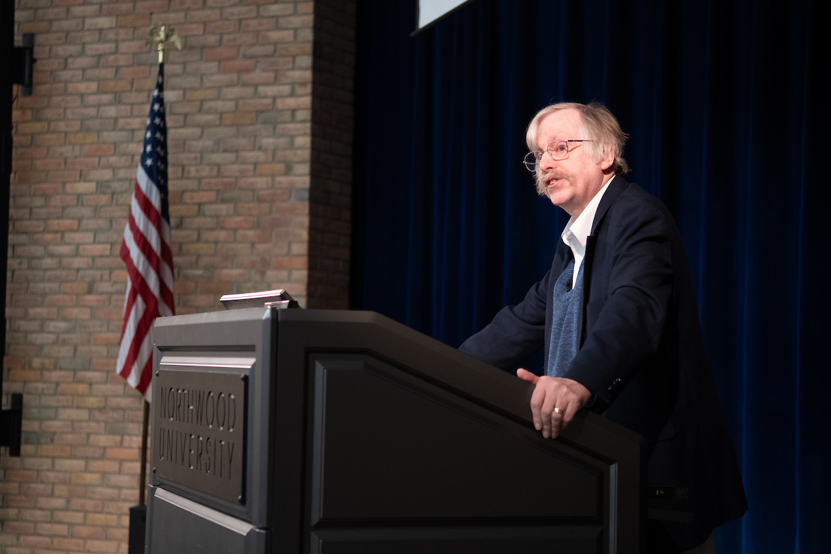 A man with glasses and a mustache speaks at a podium labeled "Northwood University," with a U.S. flag and brick wall in the background, encouraging attendees to visit campus.