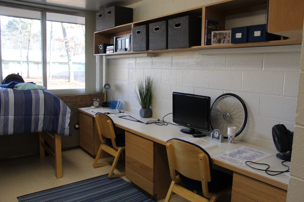 A neatly organized dorm room in Dubois and Minor Residence Halls features two study desks with chairs, 一张高架床, 仓储货架, and a window providing natural light. The desks are equipped with a computer and various desk supplies.