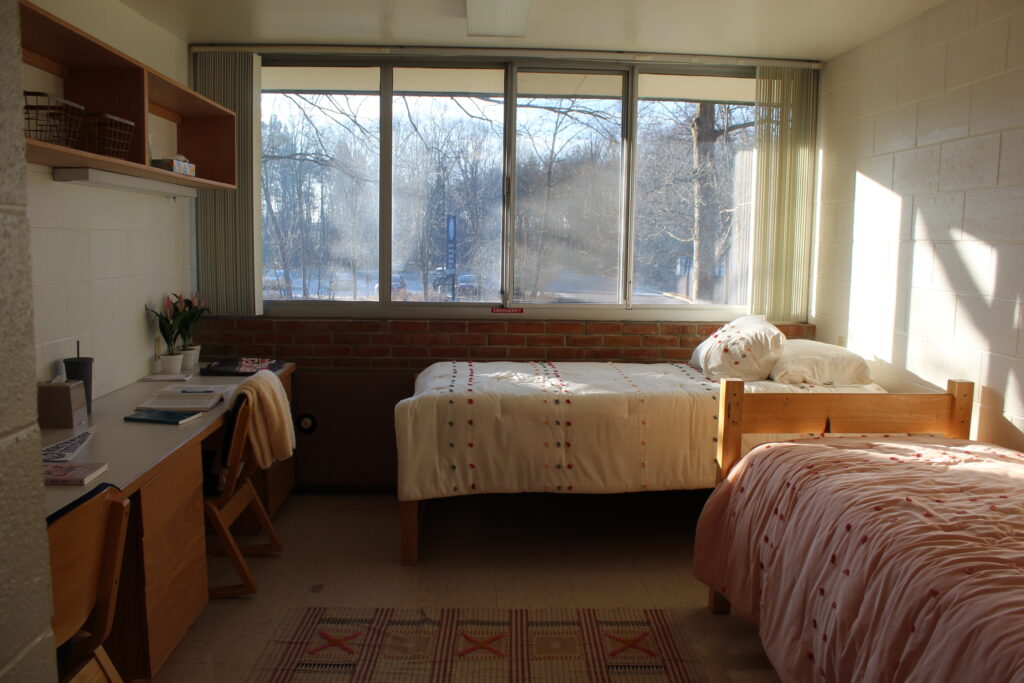 一笔可观的, sunlit dorm room in Dubois and Minor Residence Halls features two neatly made beds, a desk with chairs, a window with a scenic outdoor view, and minimal furnishings.