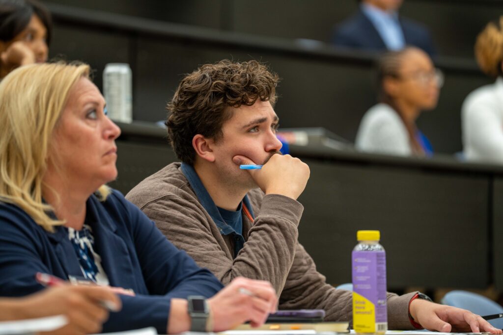 A group of people sit in a lecture hall, attentively listening. A man in the center holds a pen to his mouth, with notebooks and a drink bottle on the desk in front of him.