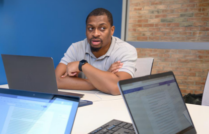 A man sits at a conference table with a laptop, speaking, while two open laptops with documents are visible in the foreground.