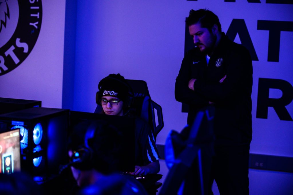 A person is seated at a computer while another person stands beside them observing. The setting appears to be an esports environment with dim lighting and visible screens.