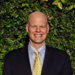 A bald man in a suit and yellow tie smiles in front of a leafy green background.