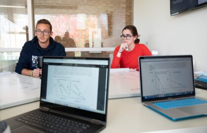 Two people sit at a conference table with laptops and papers displaying diagrams. A screen on the wall shows the day, "Thursday.