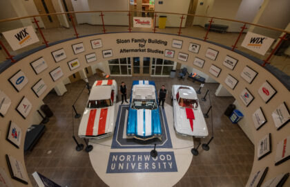 Aerial view of three classic cars displayed in the lobby of the Sloan Family Building for Aftermarket Studies at Northwood University, surrounded by sponsor logos. Four people, likely students of Automotive Aftermarket Management, stand near the cars.