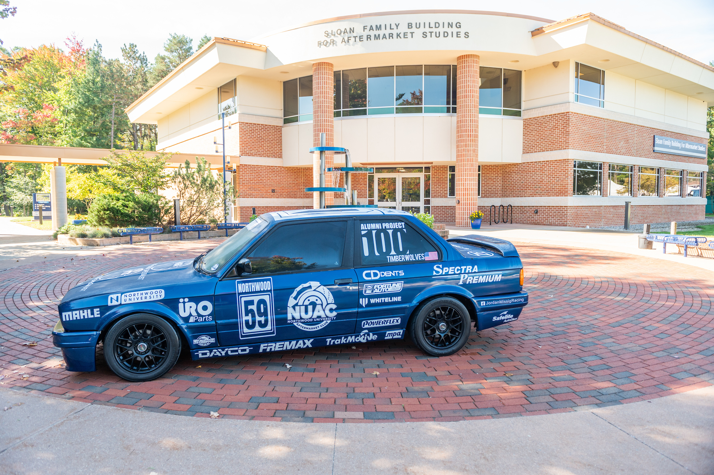 A blue race car adorned with various sponsor logos is parked in front of the Sloan Family Building, a hub for Automotive Aftermarket Management studies.