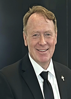 Smiling man, Dr. Michael Lee Burkhardt, CPCU, in a black suit, white shirt, and black tie with a cross pin on his lapel, standing in front of a dark background.