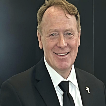 Smiling man, Dr. Michael Lee Burkhardt, CPCU, in a black suit, white shirt, and black tie with a cross pin on his lapel, standing in front of a dark background.