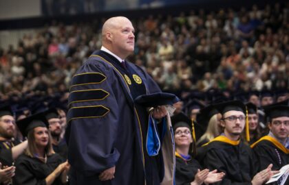 A Doctor of Business Administration in academic regalia stands proudly holding a hood at a graduation ceremony, surrounded by seated applauding graduates.