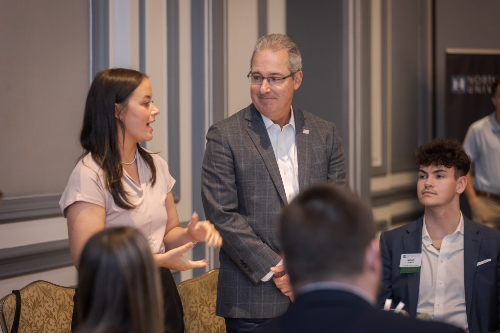 A woman stands and speaks to a man in a suit and glasses, discussing cybersecurity management, while a seated man listens attentively in the formal setting.