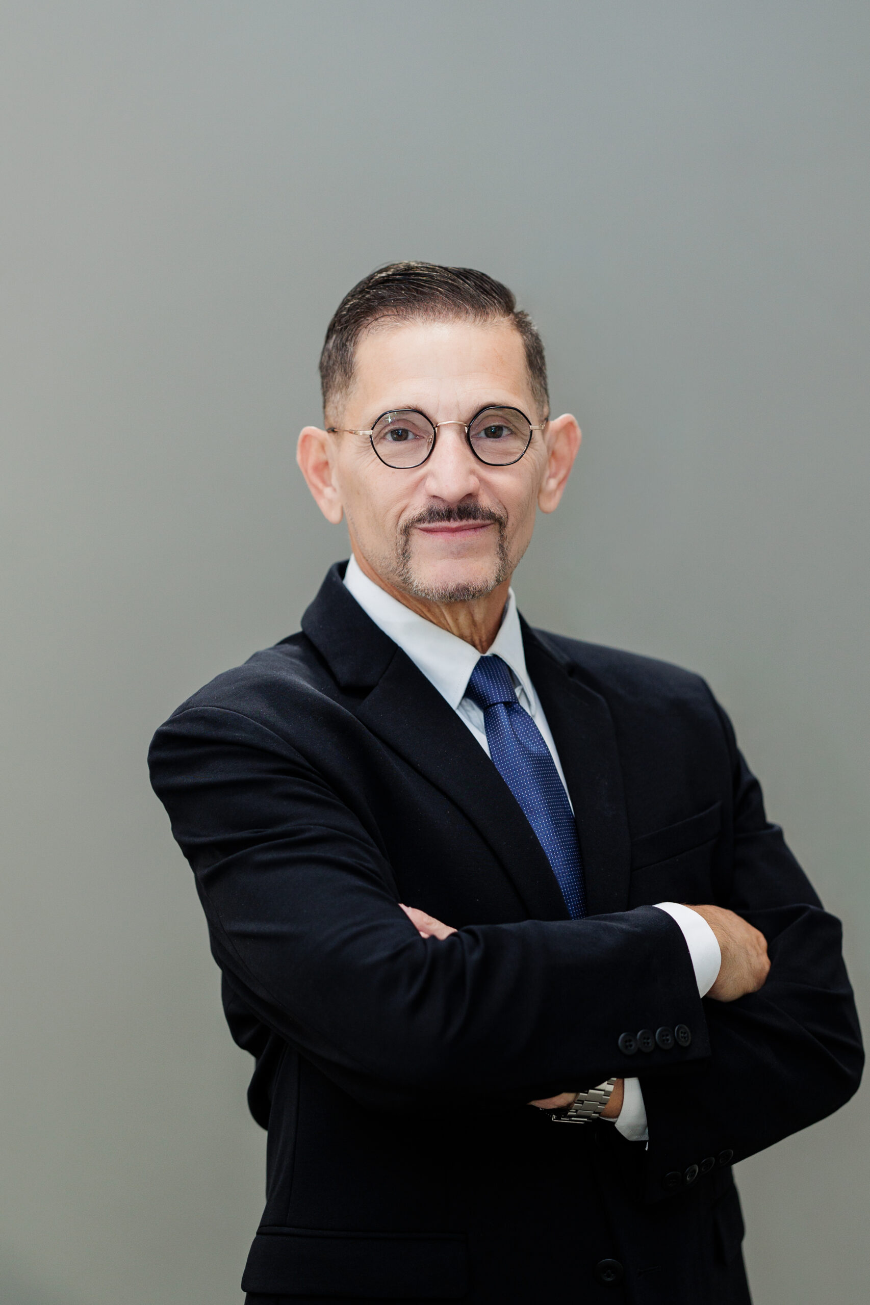 Scott Warner, a middle-aged man with a mustache and glasses, wearing a black suit, white shirt, and blue tie, posed with his arms crossed.