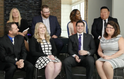 A group of nine people dressed in business attire, including a Doctor of Business Administration, are seated together indoors, smiling and chatting.