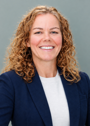 Dr. Stephanie VonFintel, a woman with curly light brown hair, wearing a navy blazer and white top, smiles in front of a plain gray background.