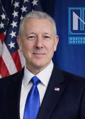 Dr. Alex Tokarev, in a suit and blue tie, stands in front of American flags and a Northwood University logo.