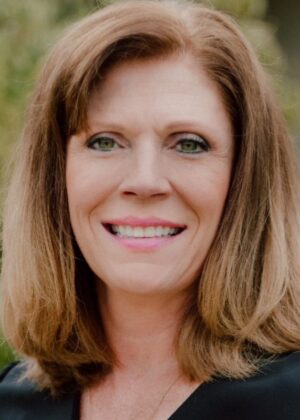 A woman with shoulder-length light brown hair and green eyes smiles at the camera, wearing a black top. The background of slightly blurred greenery adds a calm touch, reflecting her expertise in cybersecurity management.