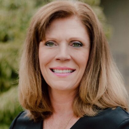 A woman with shoulder-length light brown hair and green eyes smiles at the camera, wearing a black top. The background of slightly blurred greenery adds a calm touch, reflecting her expertise in cybersecurity management.