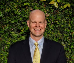 A smiling bald man in a suit and yellow tie stands in front of a leafy green background.