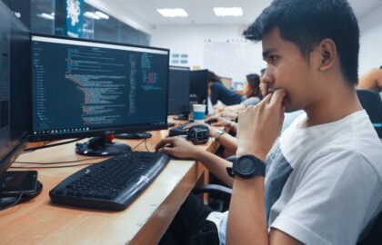 A person sits at a desk in an office, focused on a computer monitor displaying lines of code, embodying the spirit of computer science. With a keyboard and mouse in front, several other individuals work diligently in the background.