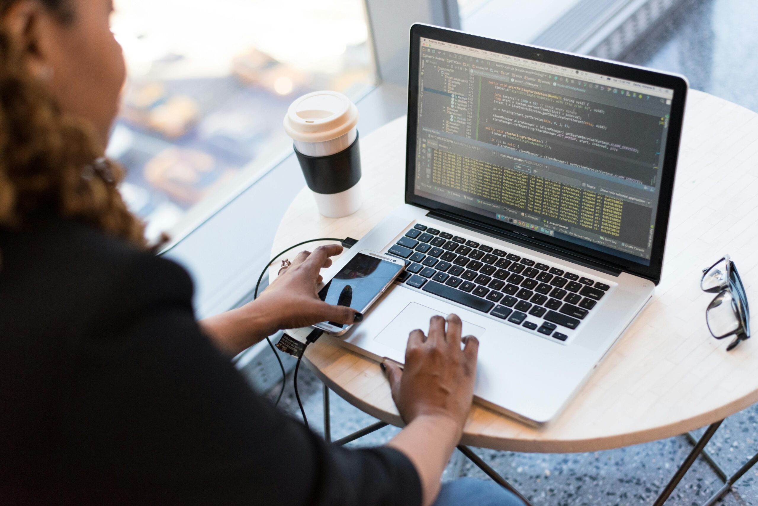 A person sits at a round table using a laptop with code on the screen, likely focused on cybersecurity management, with a smartphone, cables, and a disposable coffee cup nearby.