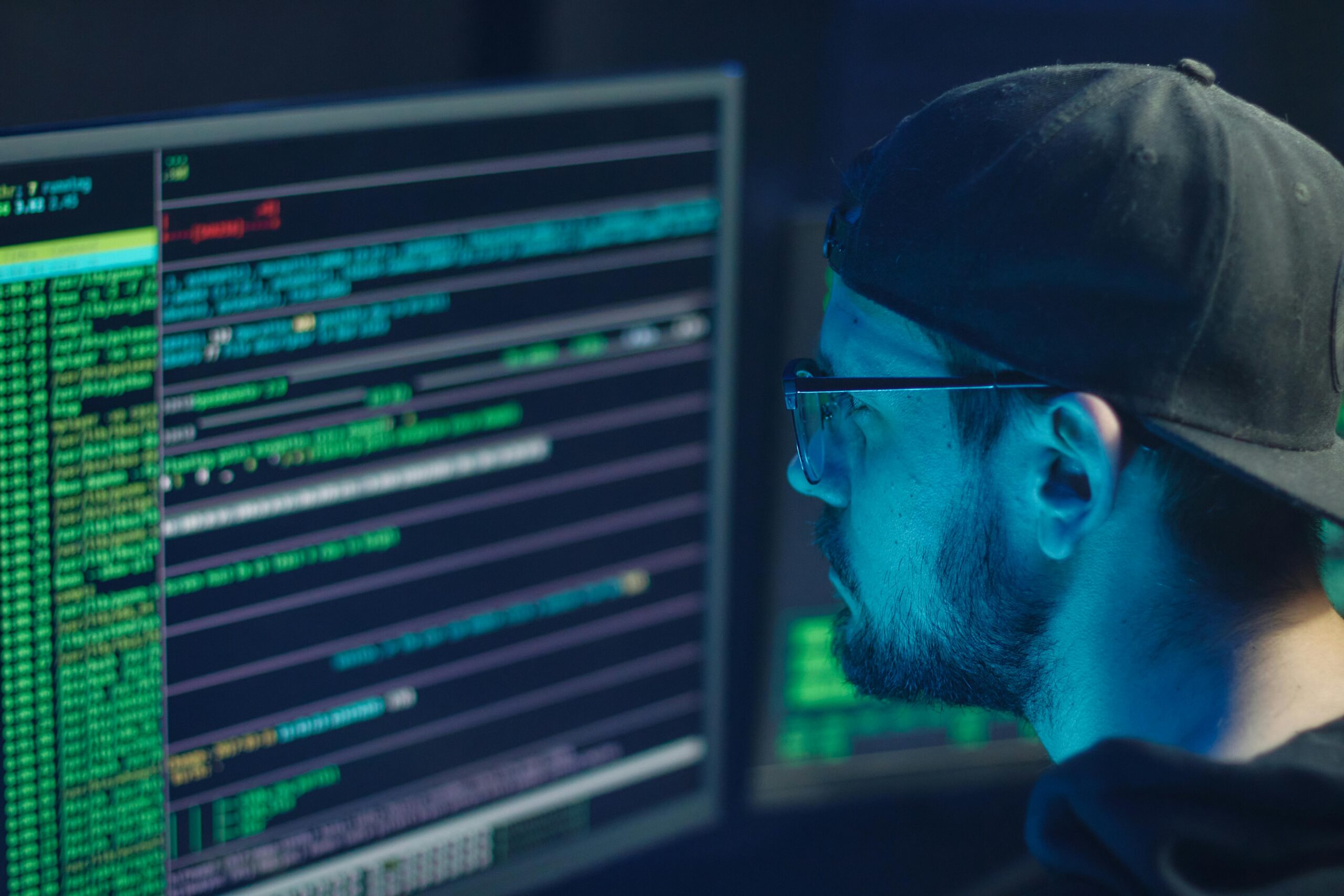 A man wearing a cap and glasses is looking at computer monitors displaying lines of code in a dimly lit room, focused on cybersecurity management.