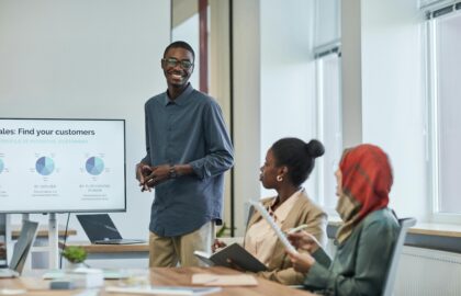 A person presenting to two colleagues in a meeting room, with a screen displaying charts about sales and customers in the background, highlights the importance of Data Analytics.