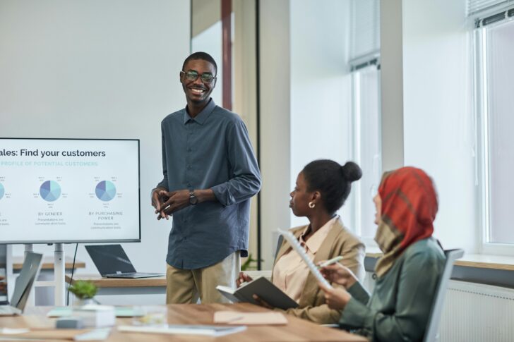 A person presenting to two colleagues in a meeting room, with a screen displaying charts about sales and customers in the background, highlights the importance of Data Analytics.