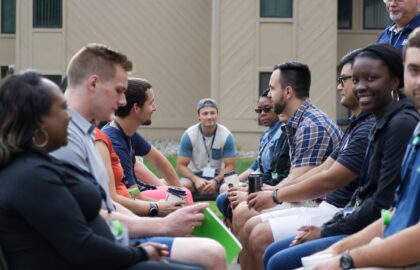 A group of people, possibly from Human Resources, is sitting outside in two rows, facing each other and engaged in conversation. Meanwhile, one person sits alone in the background, observing the group.