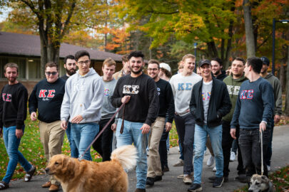 A group of young men, some wearing fraternity letters, walk outside on a college campus with two dogs. Trees with autumn foliage are in the background, creating a picturesque scene as they head towards an Interfraternity Council meeting.