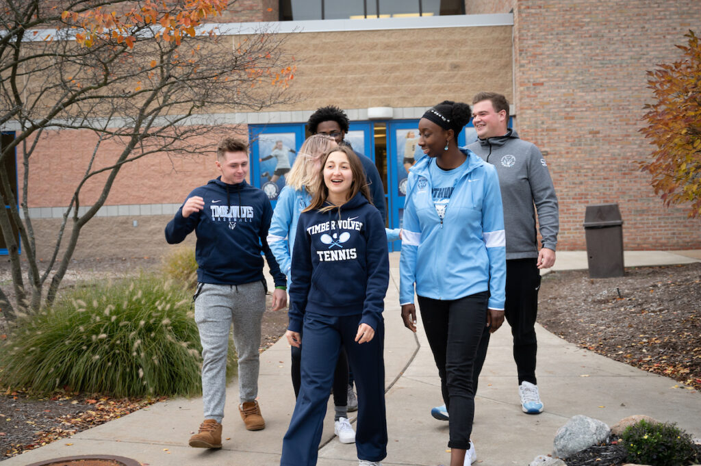 Five students dressed in school athletic clothing, walk and talk together outside a brick building on a cloudy day.
