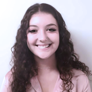 Smiling person with long, curly dark hair and a nose piercing, wearing a light pink top.