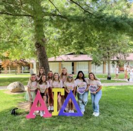 A group of women stand outdoors on grass, posing behind red, yellow, and purple Greek letters ΔΓΔ. They wear matching white t-shirts with colorful writing that proudly showcases their Alpha Gamma Delta spirit. Trees and a building are in the background.