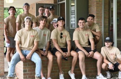 A group of young men wearing beige T-shirts and casual attire, representing Alpha Sigma Phi, pose together outside a building. Some are sitting on a low brick wall, and others are standing behind them.