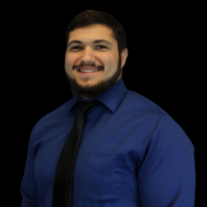 A man with a beard smiles, exemplifying confidence in his blue shirt and black tie, set against a black background.