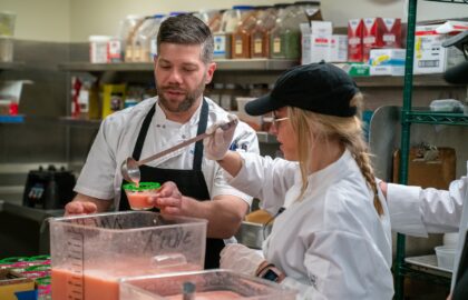 Two chefs in a kitchen, showcasing their hospitality management skills, are preparing food. One pours green sauce from a ladle into a container held by the other. Shelves brimming with cooking supplies fill the background, illustrating an efficiently managed culinary space.