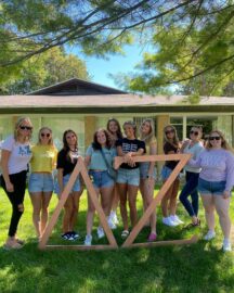 A group of eleven women stands in front of a building, posing around large wooden Delta Zeta symbols on a grassy area under a tree. They are dressed casually and smiling.