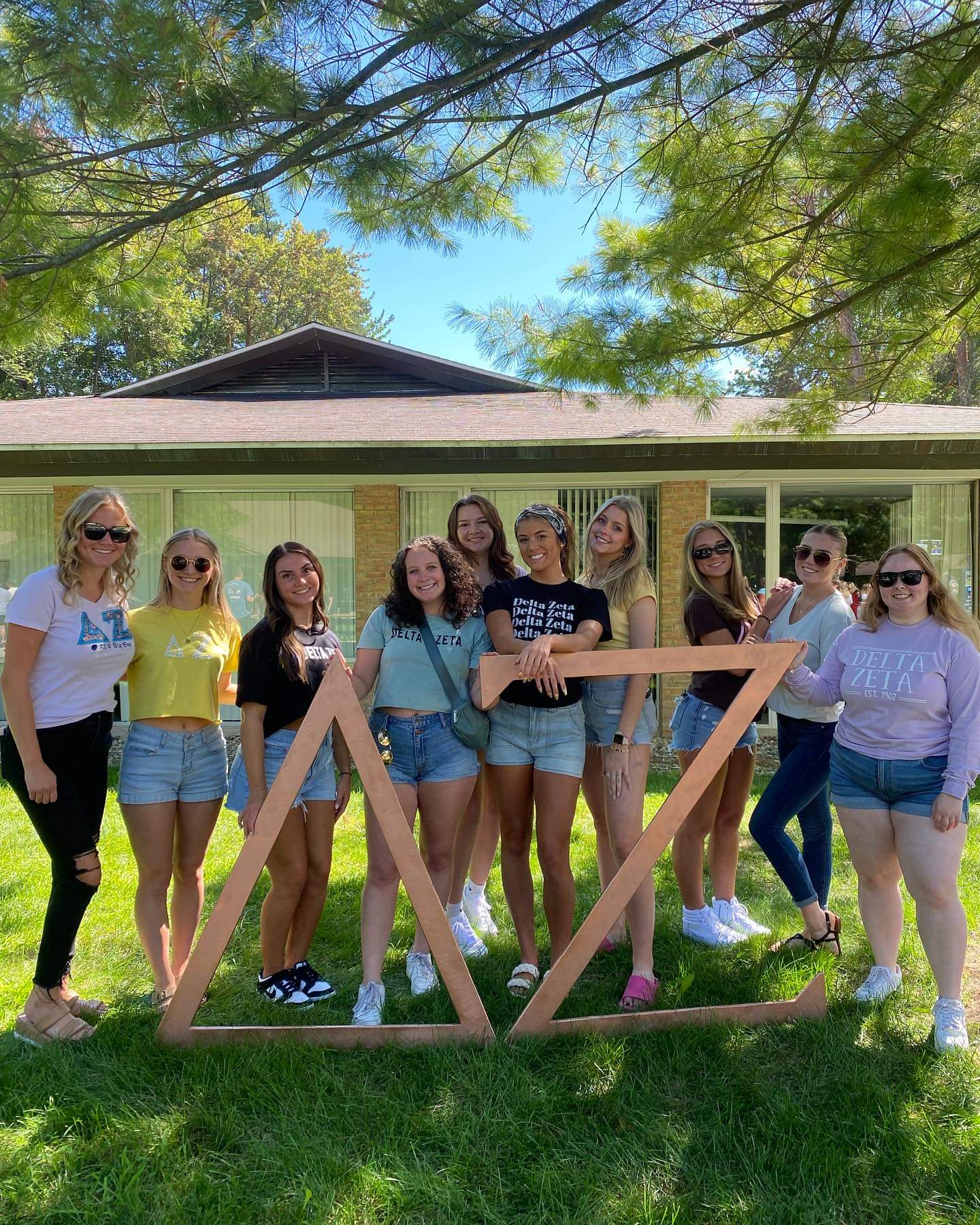 A group of eleven women stands in front of a building, posing around large wooden Delta Zeta symbols on a grassy area under a tree. They are dressed casually and smiling.