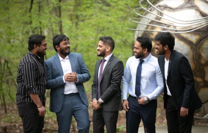 Five men in formal and casual attire, possibly discussing international business, stand outdoors, smiling and interacting against the backdrop of a large metallic sculpture.