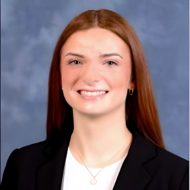 A person with long brown hair, wearing a black blazer and white top, smiles confidently against a blue background.