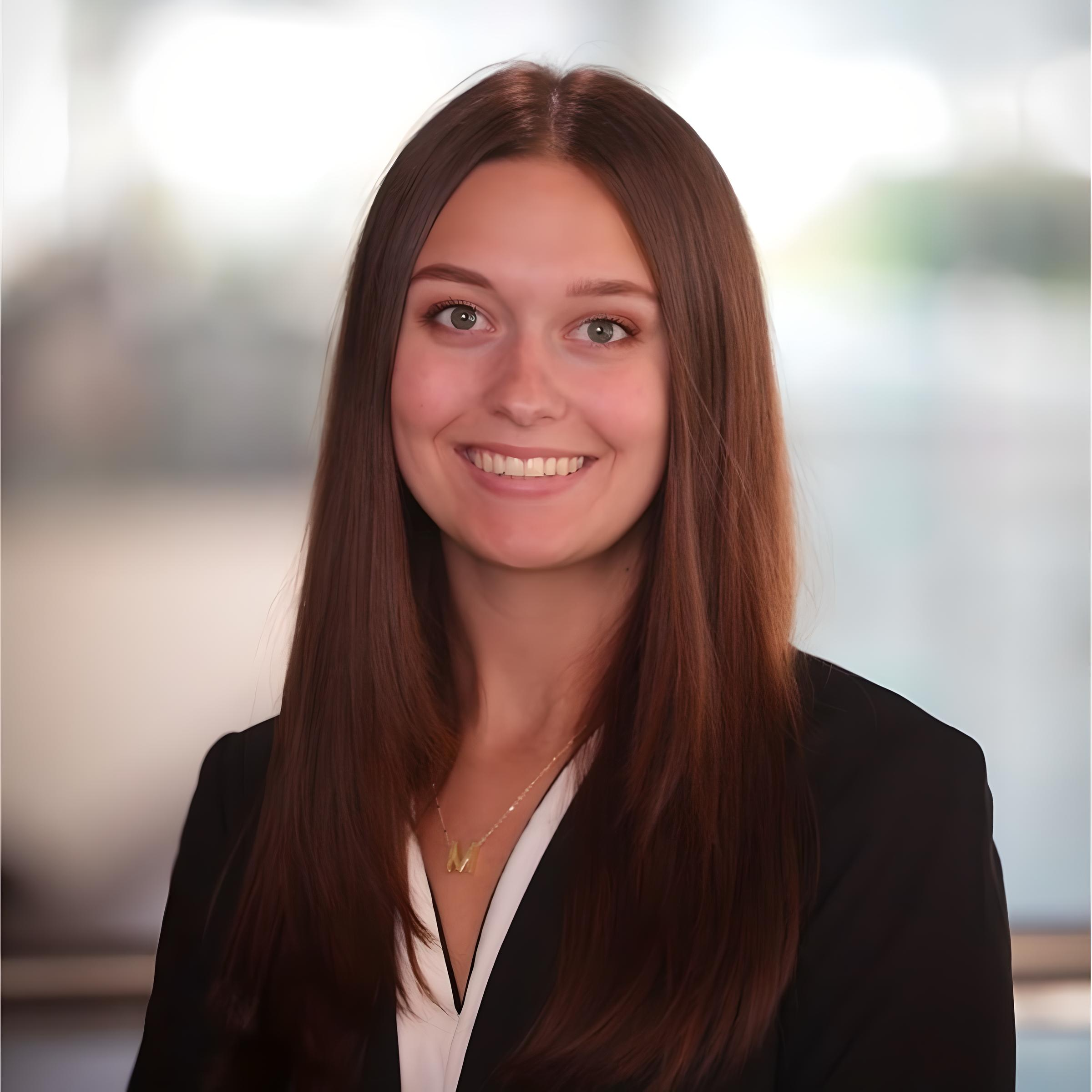 A woman with long brown hair smiles confidently, wearing a black blazer and white blouse, embodying the expertise of insurance risk management against a blurred background.