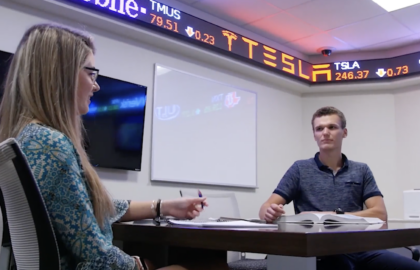 Two people are immersed in a sea of papers at a table, deeply engaged in finance discussions, while a stock ticker display rhythmically scrolls above them on the wall.