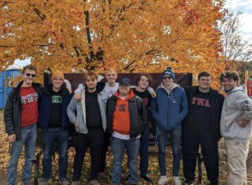 A group of nine people, some wearing clothing with the Greek letters Tau Kappa Epsilon, stand in front of a vibrant orange autumn tree, smiling at the camera.
