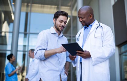 A doctor holding a tablet discusses health care management with a man in a hallway. In the background, two other people, one in scrubs, are engaged in conversation.
