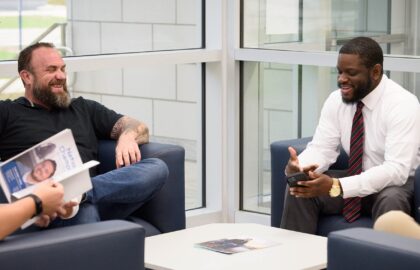 Four men are seated in a modern office lounge area, two engaged in conversation, with one reading a brochure about the Master of Business Administration program and another looking at his phone.