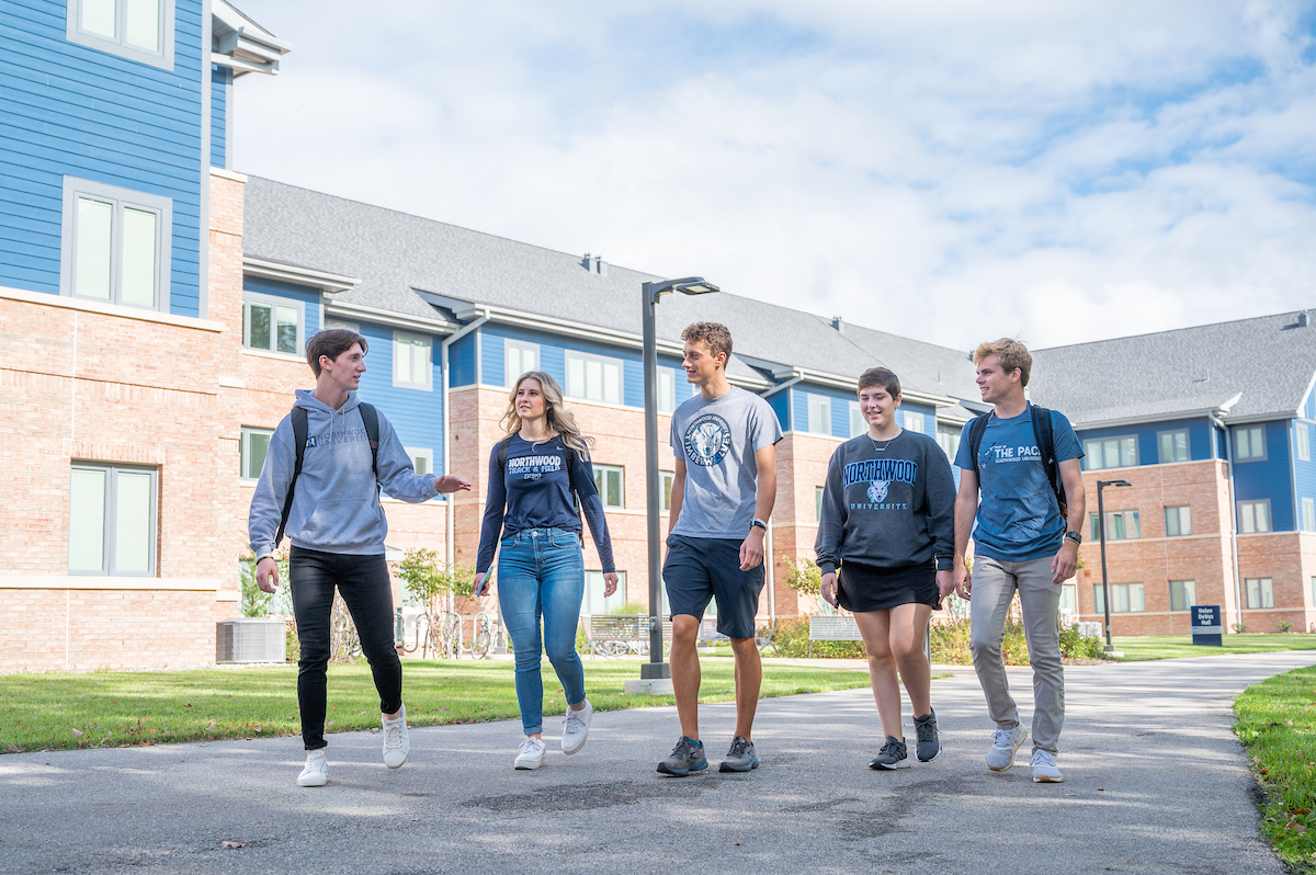 Five people walking outdoors on a college campus, with modern buildings and a cloudy sky in the background.