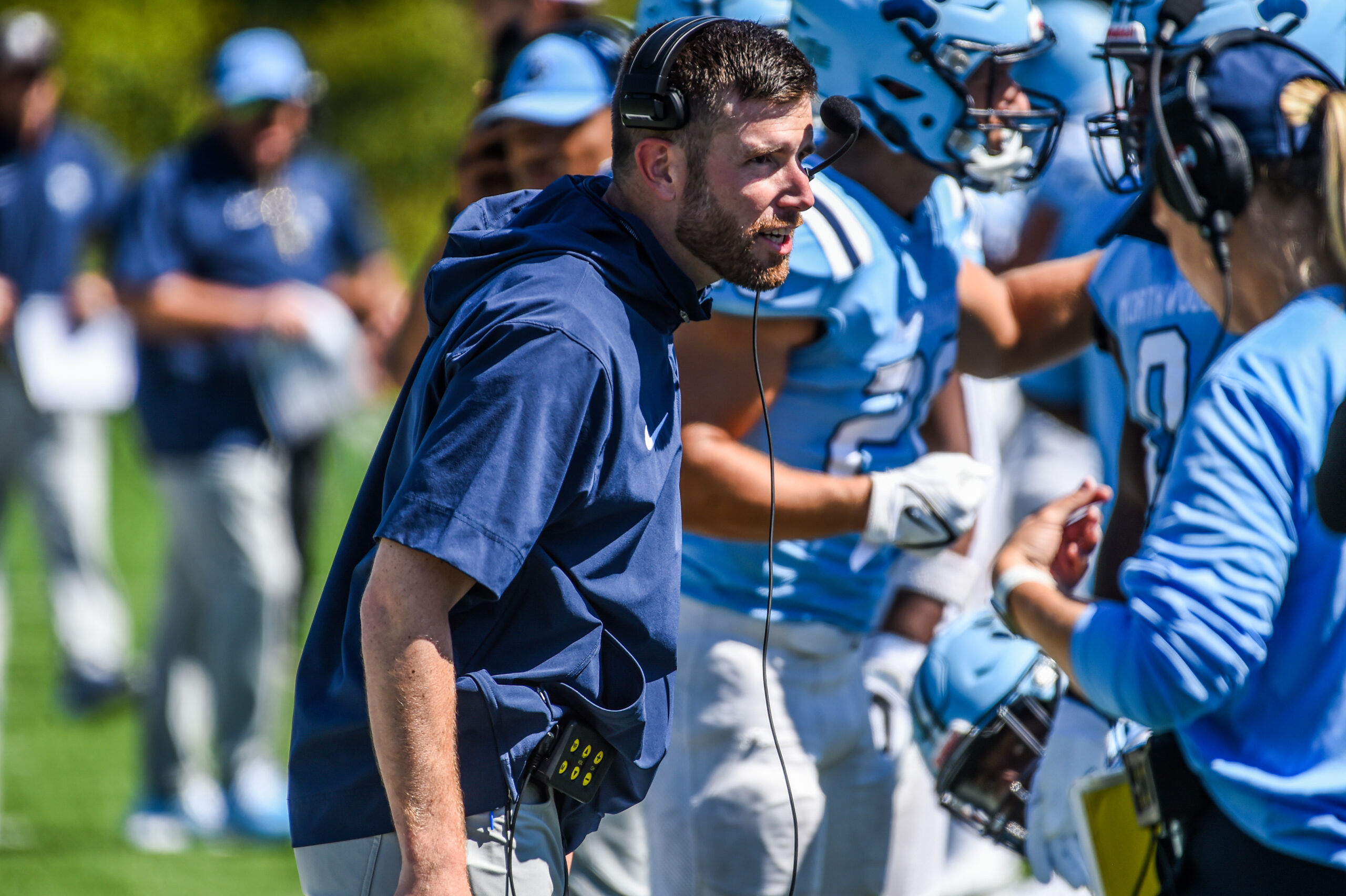 A football coach with a headset on giving directions to a group of Northwood football players and the assistant coach.