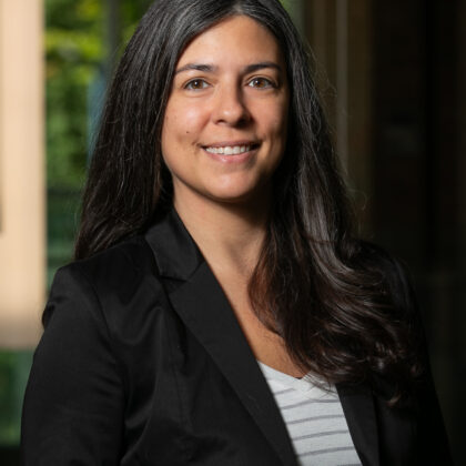 A person with long dark hair and a black blazer stands indoors, smiling.